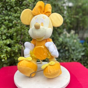 Pode incluir: Uma figura de pel&uacute;cia da Minnie Mouse em tons de amarelo e branco. A figura tem orelhas grandes e redondas, um la&ccedil;o e um vestido amarelo. Est&aacute; em uma base redonda branca, com um fundo vermelho.