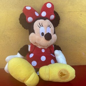 Pu&ograve; includere: Una bambola di peluche di Minnie Mouse, con un vestito rosso a pois bianchi e un fiocco abbinato. La bambola ha orecchie nere, un viso abbronzato e scarpe gialle. La bambola &egrave; seduta su una superficie rossa contro uno sfondo giallo.