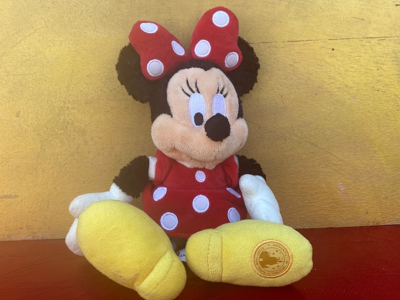 Pu&ograve; includere: Una bambola di peluche di Minnie Mouse, con un vestito rosso a pois bianchi e un fiocco abbinato. La bambola ha orecchie nere, un viso abbronzato e scarpe gialle. La bambola &egrave; seduta su una superficie rossa contro uno sfondo giallo.
