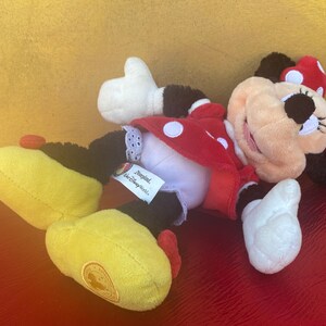 Pu&ograve; includere: Un peluche di Minnie Mouse, sdraiato su un fianco. Il giocattolo presenta un vestito rosso a pois bianchi, un fiocco abbinato e scarpe gialle. L'etichetta recita "Disneyland, Walt Disney World Parks."