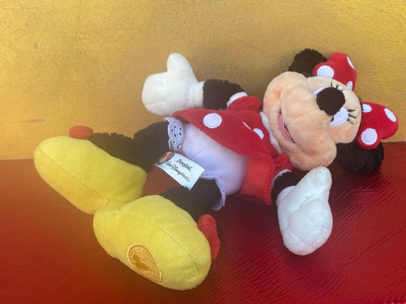 Pu&ograve; includere: Un peluche di Minnie Mouse, sdraiato su un fianco. Il giocattolo presenta un vestito rosso a pois bianchi, un fiocco abbinato e scarpe gialle. L'etichetta recita "Disneyland, Walt Disney World Parks."
