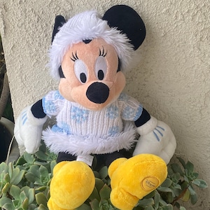K&ouml;nnte beinhalten: Ein Pl&uuml;schtier von Mickey Mouse, das einen wei&szlig;en Pullover mit blauen Schneeflocken und einem wei&szlig;en Pelzbesatz tr&auml;gt. Das Spielzeug hat einen wei&szlig;en Pelz Hut und gelbe Schuhe.