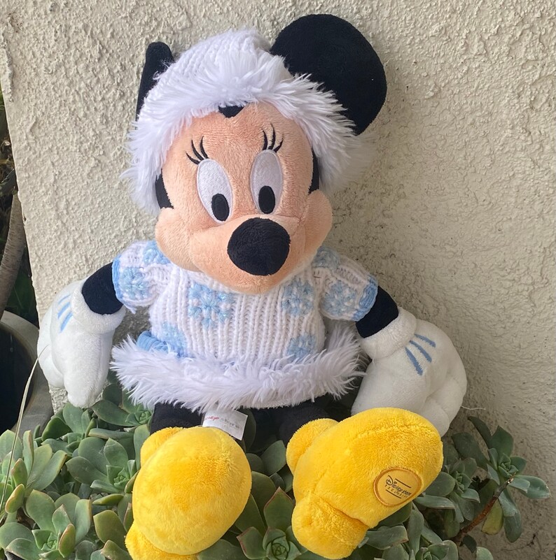 K&ouml;nnte beinhalten: Ein Pl&uuml;schtier von Mickey Mouse, das einen wei&szlig;en Pullover mit blauen Schneeflocken und einem wei&szlig;en Pelzbesatz tr&auml;gt. Das Spielzeug hat einen wei&szlig;en Pelz Hut und gelbe Schuhe.