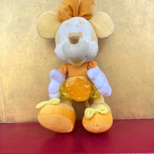Pode incluir: Uma boneca de pel&uacute;cia da Minnie Mouse em tons de laranja, amarelo e branco. A boneca tem um la&ccedil;o na cabe&ccedil;a, um vestido e sapatos. A boneca est&aacute; em uma superf&iacute;cie vermelha.