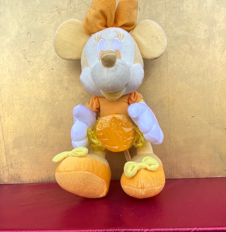 Pode incluir: Uma boneca de pel&uacute;cia da Minnie Mouse em tons de laranja, amarelo e branco. A boneca tem um la&ccedil;o na cabe&ccedil;a, um vestido e sapatos. A boneca est&aacute; em uma superf&iacute;cie vermelha.