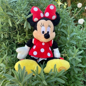 Pu&ograve; includere: Un peluche di Minnie Mouse, con un vestito rosso a pois bianchi e un fiocco abbinato. La bambola ha la pelliccia nera, un viso abbronzato e scarpe gialle. &Egrave; seduta in mezzo a un fogliame verde.