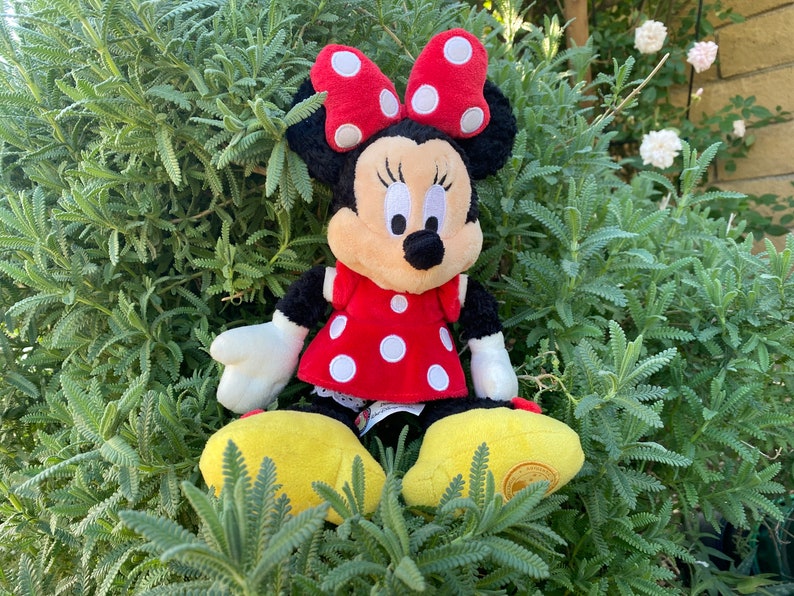 Pu&ograve; includere: Un peluche di Minnie Mouse, con un vestito rosso a pois bianchi e un fiocco abbinato. La bambola ha la pelliccia nera, un viso abbronzato e scarpe gialle. &Egrave; seduta in mezzo a un fogliame verde.