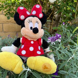 Pu&ograve; includere: Una bambola di peluche di Minnie Mouse, con un vestito rosso a pois bianchi e un fiocco abbinato. La bambola ha orecchie nere e un viso color crema. Le scarpe gialle completano il look. La bambola &egrave; seduta tra il fogliame verde.