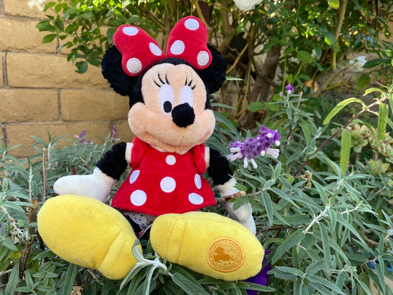 Pu&ograve; includere: Una bambola di peluche di Minnie Mouse, con un vestito rosso a pois bianchi e un fiocco abbinato. La bambola ha orecchie nere e un viso color crema. Le scarpe gialle completano il look. La bambola &egrave; seduta tra il fogliame verde.