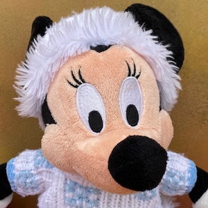 K&ouml;nnte beinhalten: Ein Pl&uuml;schtier von Minnie Mouse, das einen wei&szlig;en Pelzmantel und einen blau-wei&szlig;en Strickpullover mit Schneeflockenmuster tr&auml;gt.