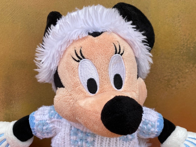 K&ouml;nnte beinhalten: Ein Pl&uuml;schtier von Minnie Mouse, das einen wei&szlig;en Pelzmantel und einen blau-wei&szlig;en Strickpullover mit Schneeflockenmuster tr&auml;gt.