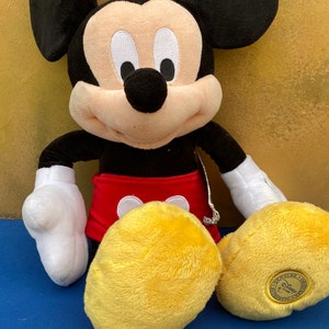 May include: A plush Mickey Mouse doll with black ears, a black nose, and a smiling face. Mickey wears red shorts, white gloves, and yellow shoes. The doll is a Disney Store product.