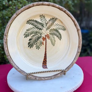 May include: A decorative plate with a palm tree design. The plate features a cream-colored background with a detailed palm tree illustration in green, brown, and orange. The rim has a bamboo-style border. The plate is displayed on a metal stand.