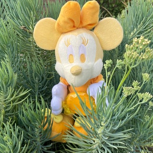 Pode incluir: Uma boneca de pel&uacute;cia Minnie Mouse amarela, com vestido e la&ccedil;o amarelos, luvas e sapatos brancos. A boneca est&aacute; rodeada por folhagem verde. A boneca tem tra&ccedil;os faciais bordados.