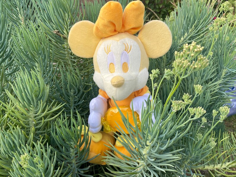 Pode incluir: Uma boneca de pel&uacute;cia Minnie Mouse amarela, com vestido e la&ccedil;o amarelos, luvas e sapatos brancos. A boneca est&aacute; rodeada por folhagem verde. A boneca tem tra&ccedil;os faciais bordados.