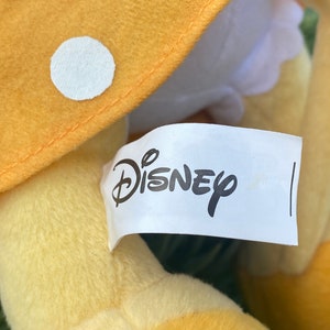 Pode incluir: Um brinquedo de pel&uacute;cia com um chap&eacute;u de cogumelo amarelo com bolinhas brancas. O logotipo da Disney est&aacute; impresso em uma etiqueta branca presa ao brinquedo. O brinquedo tem uma textura macia e fofa.