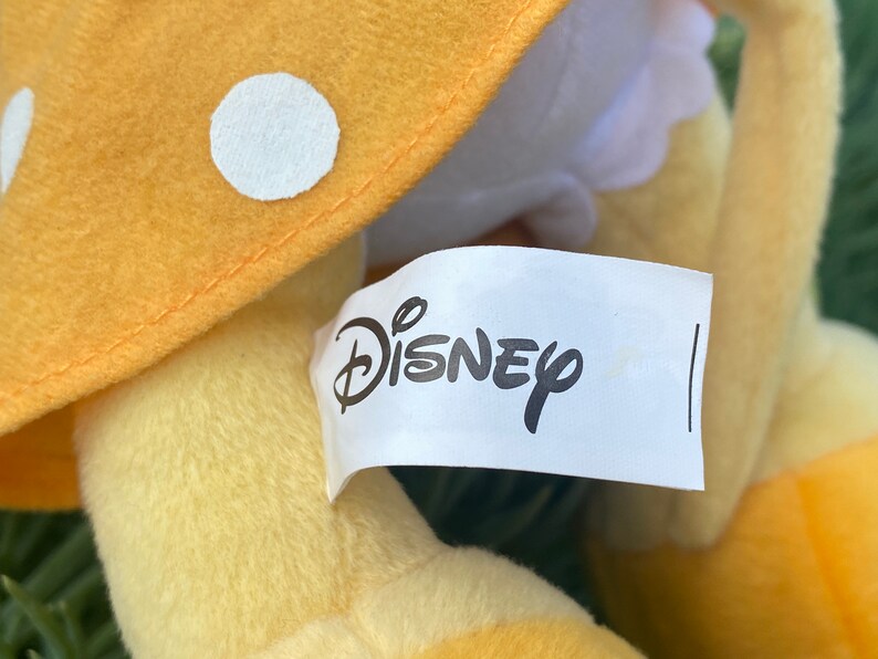 Pode incluir: Um brinquedo de pel&uacute;cia com um chap&eacute;u de cogumelo amarelo com bolinhas brancas. O logotipo da Disney est&aacute; impresso em uma etiqueta branca presa ao brinquedo. O brinquedo tem uma textura macia e fofa.