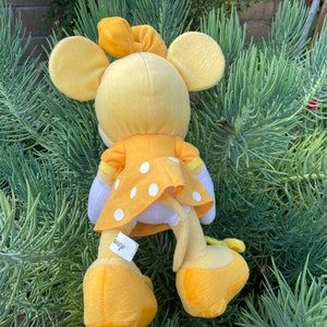 Pode incluir: Um brinquedo de pel&uacute;cia amarelo de um personagem com grandes orelhas redondas, usando um vestido amarelo com bolinhas brancas. O brinquedo tem um la&ccedil;o amarelo e est&aacute; sobre um fundo de folhagem verde. Uma etiqueta com a palavra "Disney" &eacute; vis&iacute;vel.