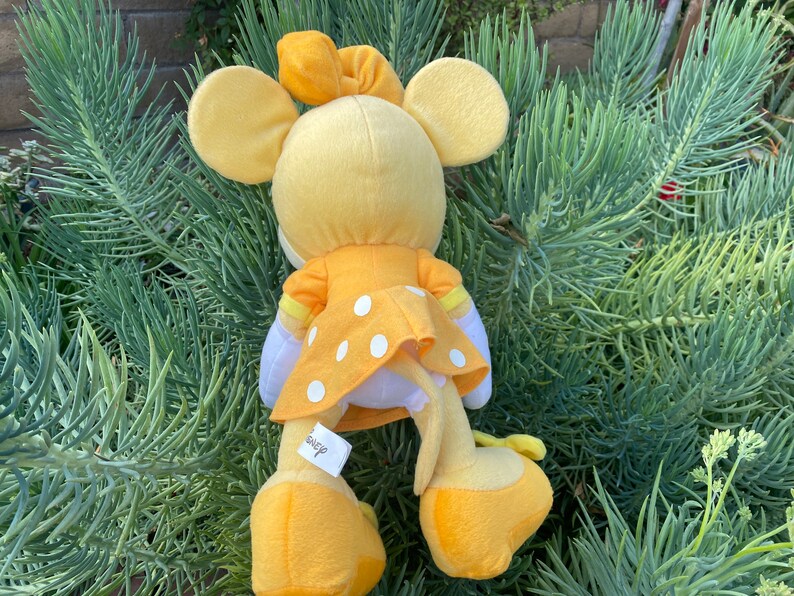 Pode incluir: Um brinquedo de pel&uacute;cia amarelo de um personagem com grandes orelhas redondas, usando um vestido amarelo com bolinhas brancas. O brinquedo tem um la&ccedil;o amarelo e est&aacute; sobre um fundo de folhagem verde. Uma etiqueta com a palavra "Disney" &eacute; vis&iacute;vel.