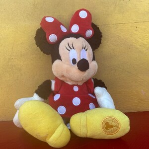 Pu&ograve; includere: Un peluche di Minnie Mouse, con un vestito rosso a pois bianchi e un fiocco abbinato. Il giocattolo ha orecchie nere, un viso sorridente e piedi gialli. Il peluche &egrave; seduto su una superficie rossa.