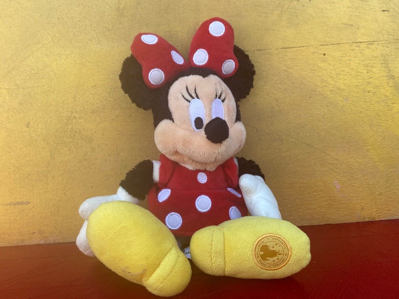 Pu&ograve; includere: Un peluche di Minnie Mouse, con un vestito rosso a pois bianchi e un fiocco abbinato. Il giocattolo ha orecchie nere, un viso sorridente e piedi gialli. Il peluche &egrave; seduto su una superficie rossa.
