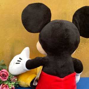 May include: A plush Mickey Mouse toy, viewed from the back, with large black ears, a black head, and a red pair of shorts. The toy's white gloved hand is extended towards a yellow object. Pink roses are in the foreground.