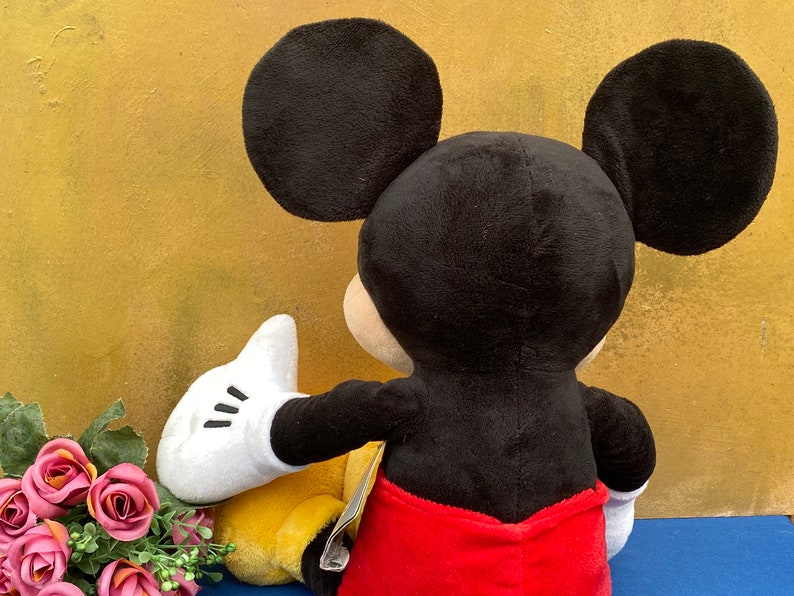 May include: A plush Mickey Mouse toy, viewed from the back, with large black ears, a black head, and a red pair of shorts. The toy's white gloved hand is extended towards a yellow object. Pink roses are in the foreground.
