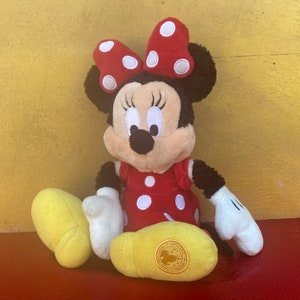 Pu&ograve; includere: Una bambola di peluche di Minnie Mouse, con un vestito rosso a pois bianchi e un fiocco abbinato. La bambola ha orecchie nere, un naso nero e guanti bianchi. Sono visibili scarpe gialle. Lo sfondo &egrave; una superficie gialla.