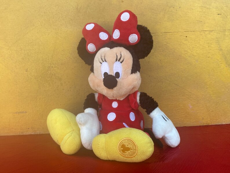 Pu&ograve; includere: Una bambola di peluche di Minnie Mouse, con un vestito rosso a pois bianchi e un fiocco abbinato. La bambola ha orecchie nere, un naso nero e guanti bianchi. Sono visibili scarpe gialle. Lo sfondo &egrave; una superficie gialla.