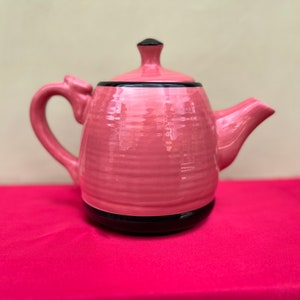 Könnte beinhalten: Eine rosa Keramik-Teekanne mit schwarzem Rand und Deckel. Die Teekanne hat einen gebogenen Griff und Ausguss sowie einen kleinen Knopf auf dem Deckel. Die Teekanne steht auf einer rosa Oberfläche.