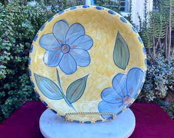 May include: A hand-painted ceramic serving plate with a yellow background and blue floral design. The plate has a scalloped edge with blue and brown accents. The flowers have blue petals and brown centers, with green leaves.