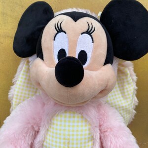 May include: Pink and yellow plush toy of a cartoon mouse with large black ears and a gingham bow. The mouse has a black nose and white eyes with eyelashes.
