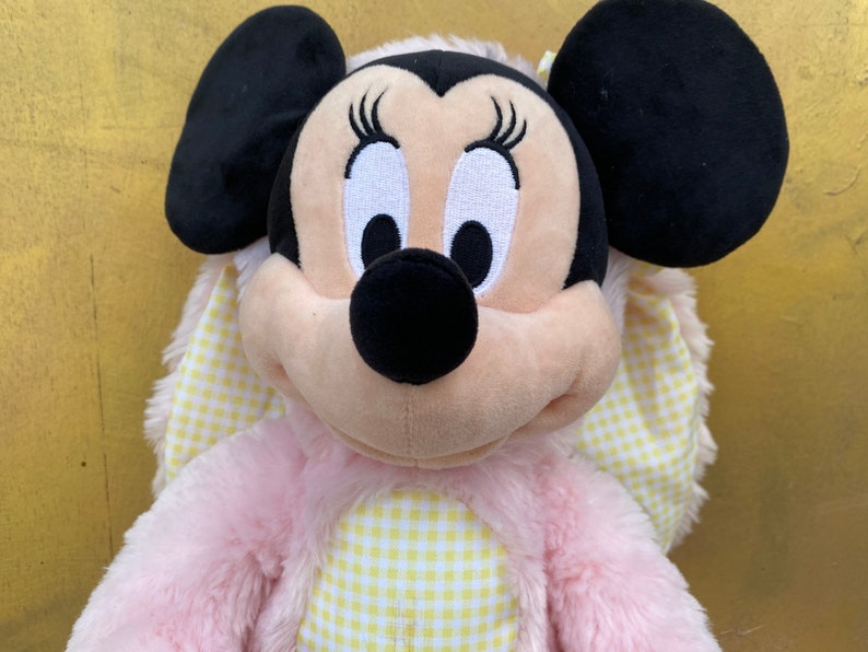 May include: Pink and yellow plush toy of a cartoon mouse with large black ears and a gingham bow. The mouse has a black nose and white eyes with eyelashes.