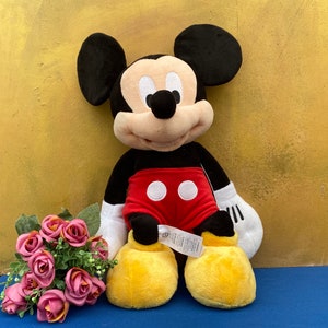 May include: A plush Mickey Mouse doll, featuring black ears, a black body, red shorts with white buttons, and yellow shoes. The doll has a cream-colored face and white gloves. A bouquet of pink roses rests nearby.