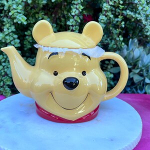 May include: A yellow ceramic teapot shaped like Winnie the Pooh's face, with a black nose, smiling mouth, and small black eyes. The teapot has a red band around the base and a white frilly lid.