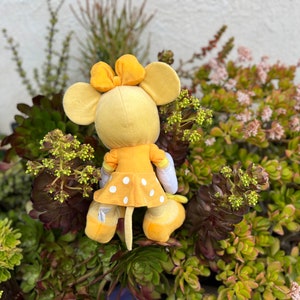 Pode incluir: Um brinquedo de pel&uacute;cia amarelo em forma de rato, com la&ccedil;o, orelhas e vestido de bolinhas. O brinquedo est&aacute; rodeado por suculentas e plantas verdes e roxas, criando um contraste vibrante.