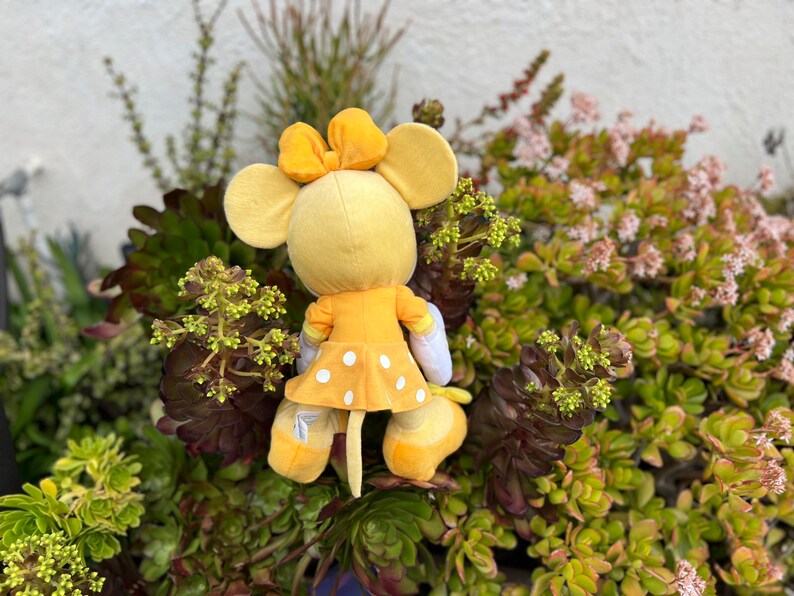 Pode incluir: Um brinquedo de pel&uacute;cia amarelo em forma de rato, com la&ccedil;o, orelhas e vestido de bolinhas. O brinquedo est&aacute; rodeado por suculentas e plantas verdes e roxas, criando um contraste vibrante.