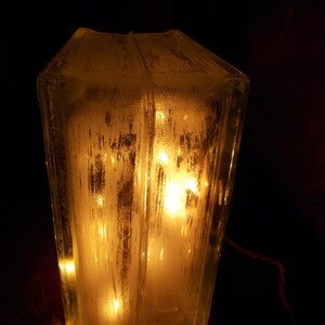Peut inclure: Une lampe en verre transparent avec une surface textur&eacute;e, &eacute;clair&eacute;e par des lumi&egrave;res blanches chaudes &agrave; l'int&eacute;rieur. La lampe est pos&eacute;e sur une surface en bois.