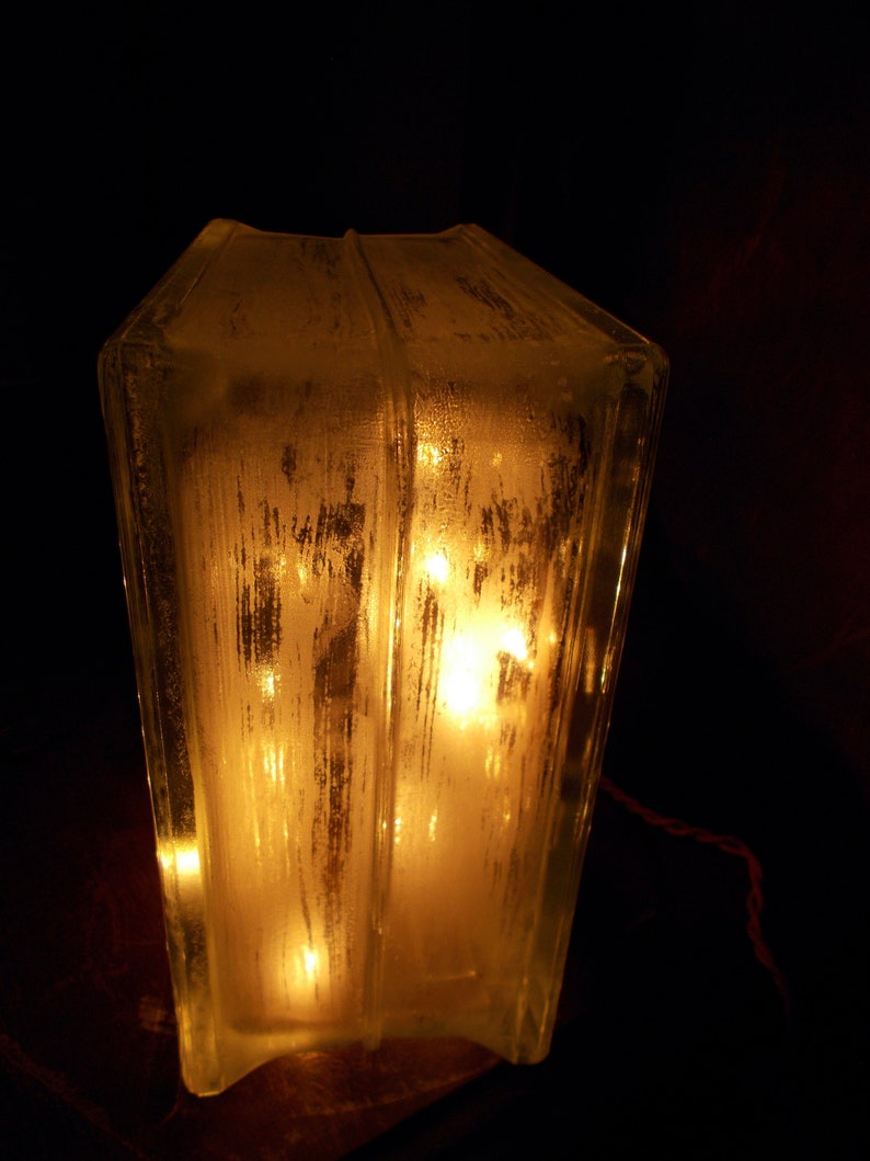 Peut inclure: Une lampe en verre transparent avec une surface textur&eacute;e, &eacute;clair&eacute;e par des lumi&egrave;res blanches chaudes &agrave; l'int&eacute;rieur. La lampe est pos&eacute;e sur une surface en bois.