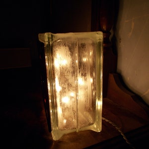 Peut inclure: Un bloc de verre transparent avec une guirlande lumineuse &agrave; l'int&eacute;rieur, illuminant le bloc de l'int&eacute;rieur. Le bloc est pos&eacute; sur une surface en bois fonc&eacute;.