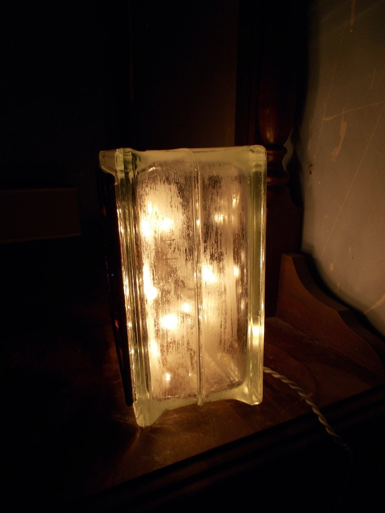 Peut inclure: Un bloc de verre transparent avec une guirlande lumineuse &agrave; l'int&eacute;rieur, illuminant le bloc de l'int&eacute;rieur. Le bloc est pos&eacute; sur une surface en bois fonc&eacute;.