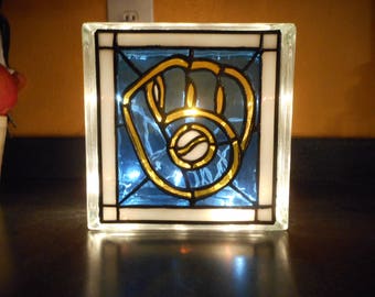 May include: A glass block with a stained glass design of a baseball glove and ball. The glove is gold and the ball is white. The background is blue.