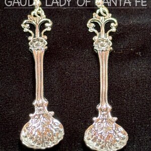 May include: A pair of silver-toned spoon earrings with intricate floral and scrollwork detailing. The earrings have a vintage aesthetic, with curved ear wires and a decorative top. The text "GAUDY LADY OF SANTA FE" is printed above the earrings.