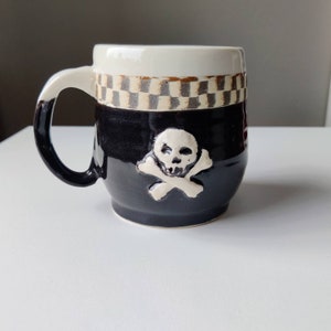 May include: A black ceramic mug with a white skull and crossbones design. The mug has a checkered pattern around the top.