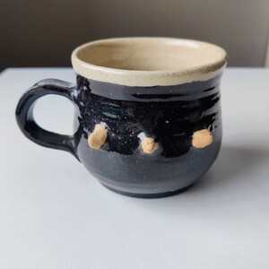 May include: A black ceramic mug with a cream-colored rim and three small, beige bumps on the side.