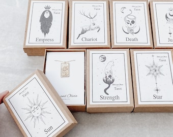 May include: A set of ten small, brown cardboard boxes with gold lettering and illustrations. Each box features a different tarot card image, including Empress, Chariot, Death, Judgement, Sun, Strength, Star, and more. The boxes are arranged in two rows, with a necklace in a white box in the center.