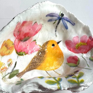 May include: A decorative, white, seashell-shaped dish with a painted design. The design features a yellow bird, pink and yellow flowers, and a blue dragonfly. The dish is likely used for holding small items or as a decorative piece.