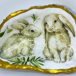May include: A decorative oyster shell dish featuring two watercolor-style rabbits in shades of beige and green. The shell has a gold-painted rim and is filled with a white background and green foliage.
