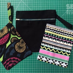May include: Three zippered pouches in various sizes and patterns. One is solid black with a gold zipper, another features a colorful geometric design, and the third has a black, white, pink, and yellow tribal pattern. Each pouch has a matching fabric wrist strap.