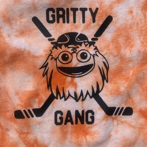 May include: Orange and white tie-dye t-shirt with a black and white graphic of a furry orange creature wearing a hockey helmet and holding two hockey sticks. The text "Gritty Gang" is printed below the creature.